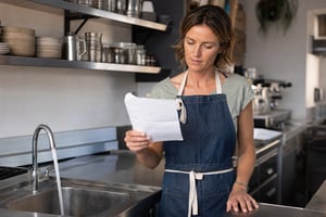 Cafe owner in an apron reading a business water bill beside a running kitchen tap